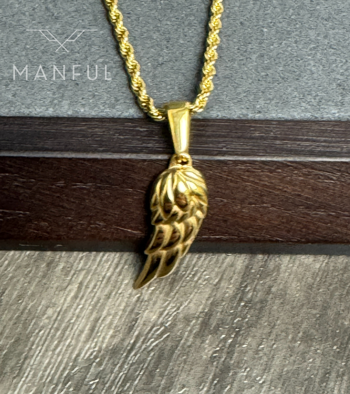 Angel Wing Pendant Necklace Gold, propped against a stone and dark wood ledge, chain draped to one side