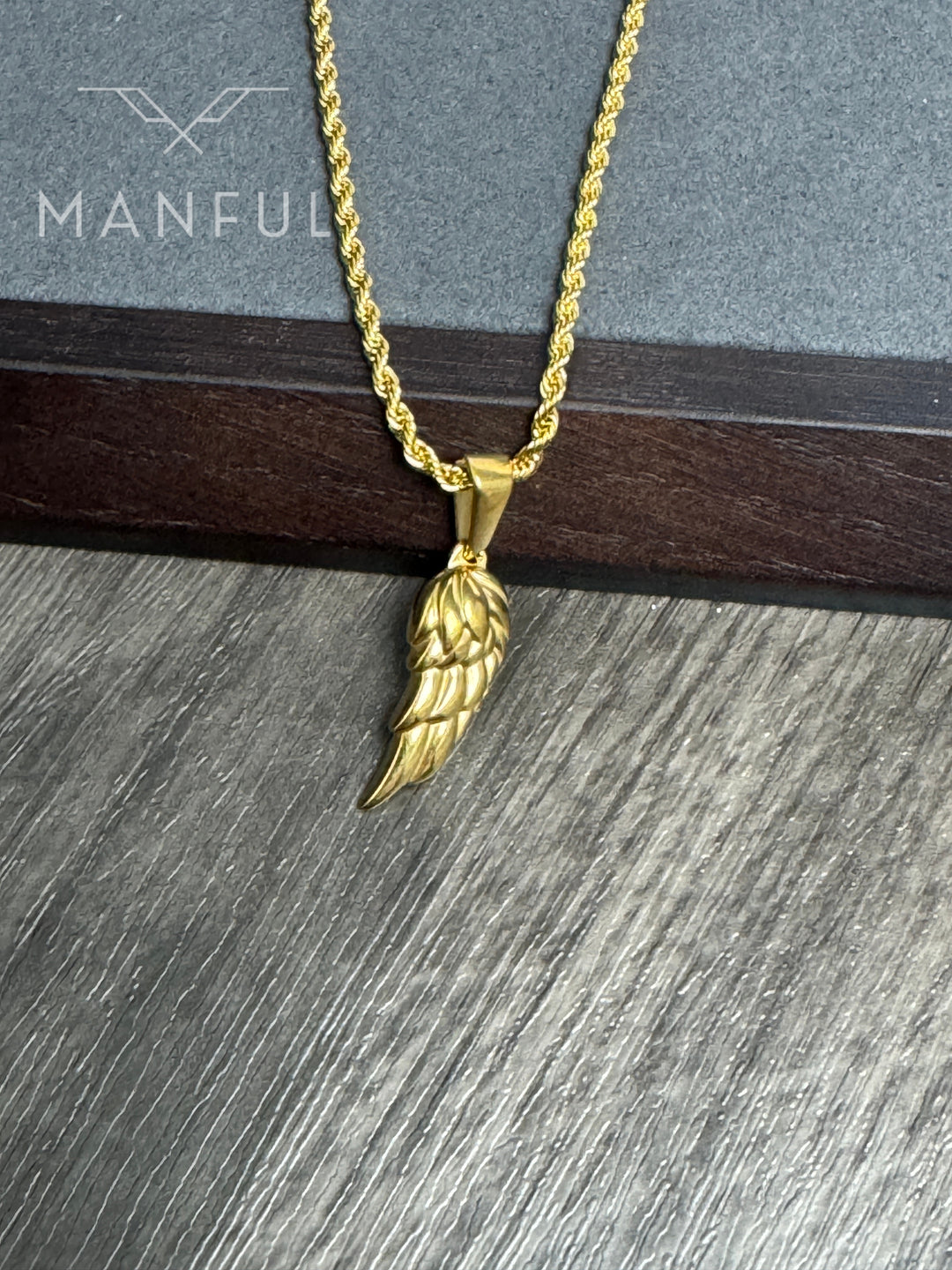 Angel Wing Pendant Necklace Gold, close-up against a stone and wood ledge showing the wing's feather detail