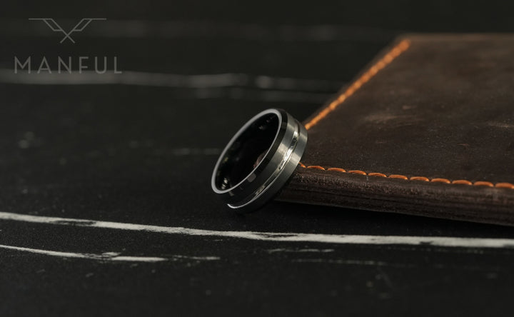 Black and Silver Tungsten Ring, propped against a leather-bound book with orange stitching