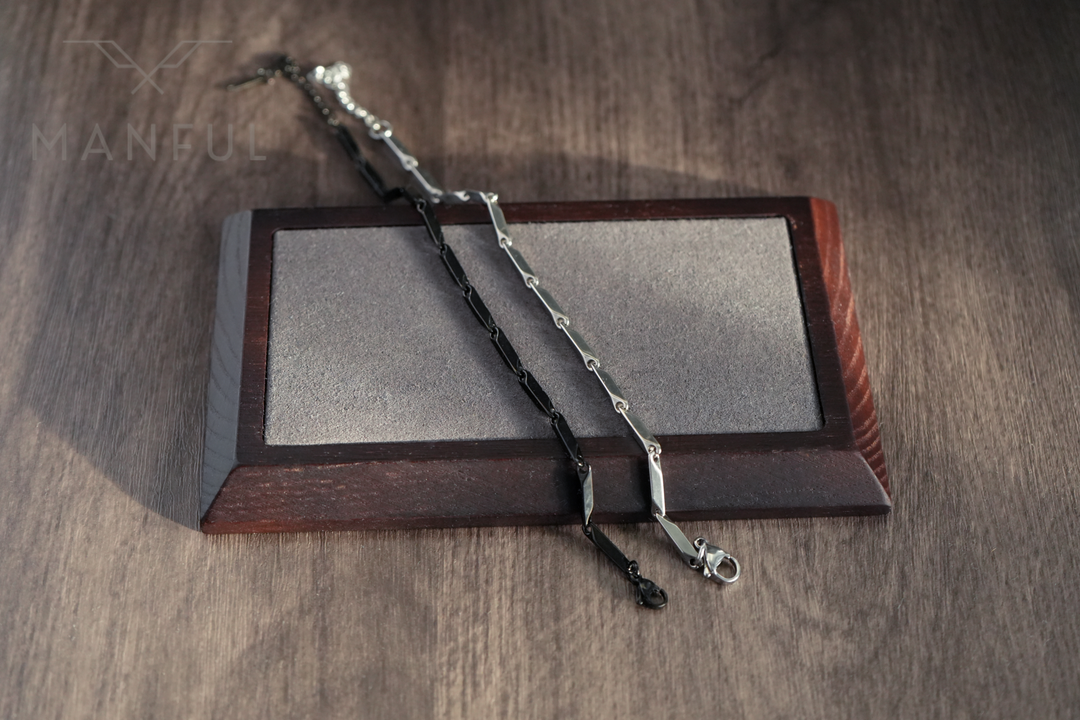 Block Chain Bracelet Black, draped over a wooden stand beside a silver chain bracelet
