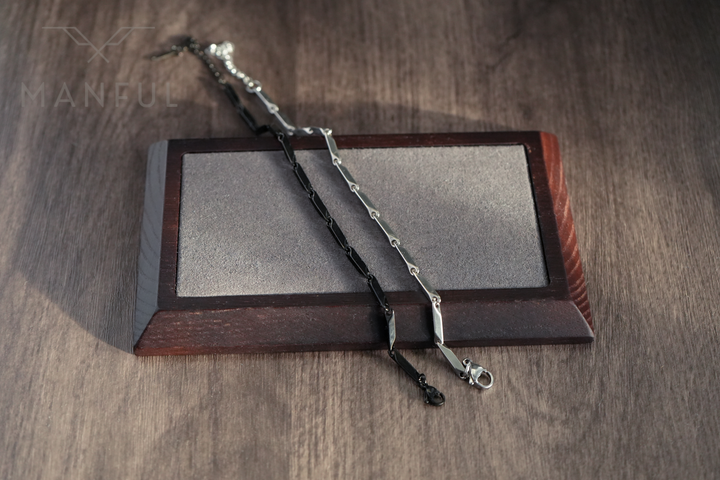 Block Chain Bracelet Black, draped over a wooden stand beside a silver chain bracelet