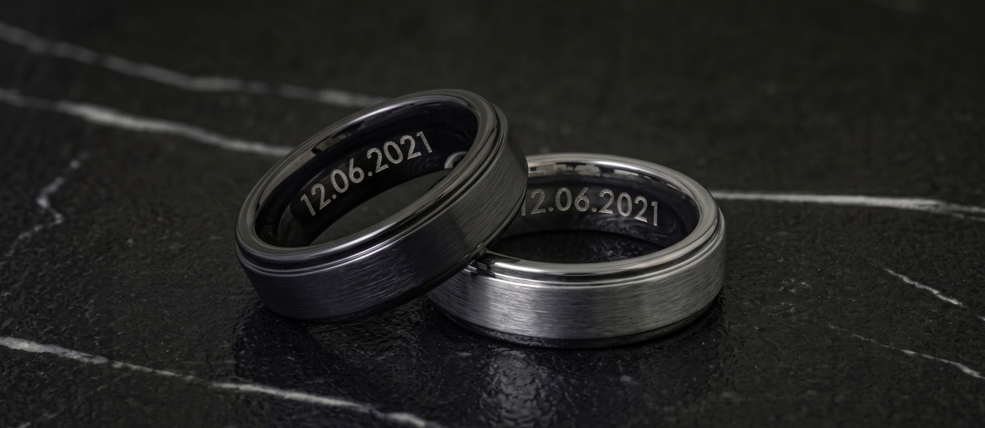 Brushed Tungsten Bands in black and silver on dark marble, each engraved inside with the date 12.06.2021