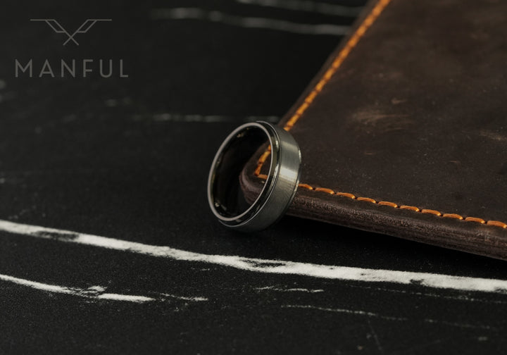 Brushed Tungsten Band Gunmetal, resting against a leather book with orange stitching on dark slate