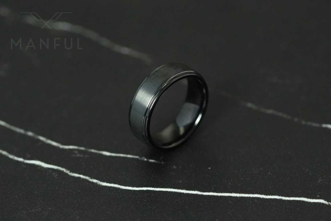 Brushed Tungsten Band Black, resting at an angle on a dark cracked stone slab, tilted to the right