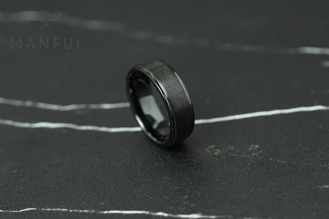 Brushed Tungsten Band Black, resting at an angle on a dark cracked stone slab, tilted to the left
