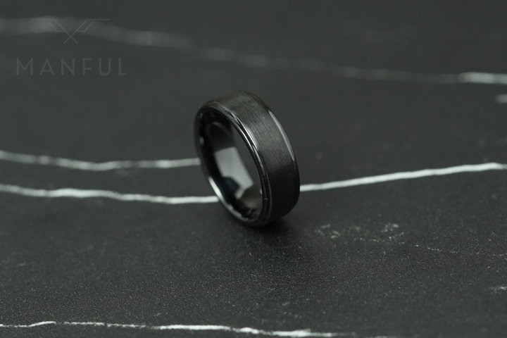 Brushed Tungsten Band Black, resting at an angle on a dark cracked stone slab, tilted to the left