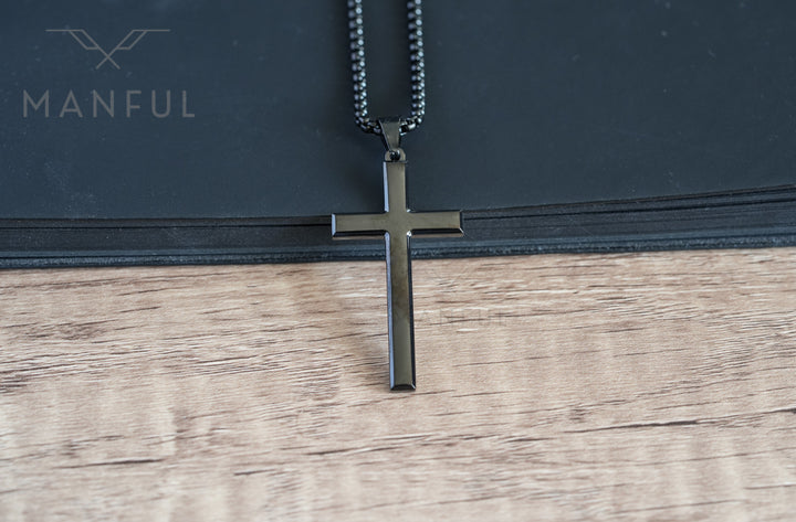 Cross Pendant Necklace Black, hanging against a black book edge on a wood surface