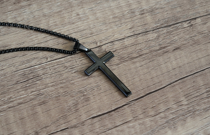Cross Pendant Necklace Black, lying flat on a wood surface with the chain coiled beside it