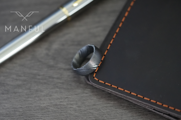 Damascus Steel Band, propped against an orange-stitched leather book beside a pen