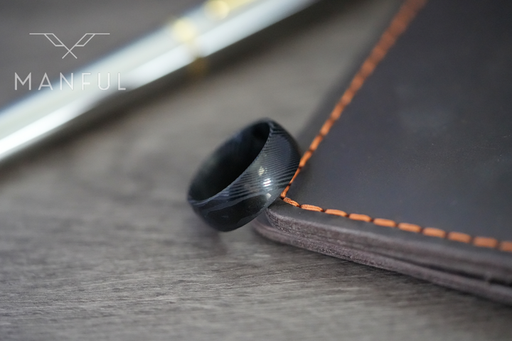 Damascus Steel Band Black, leaning against a dark book with orange stitching on wood