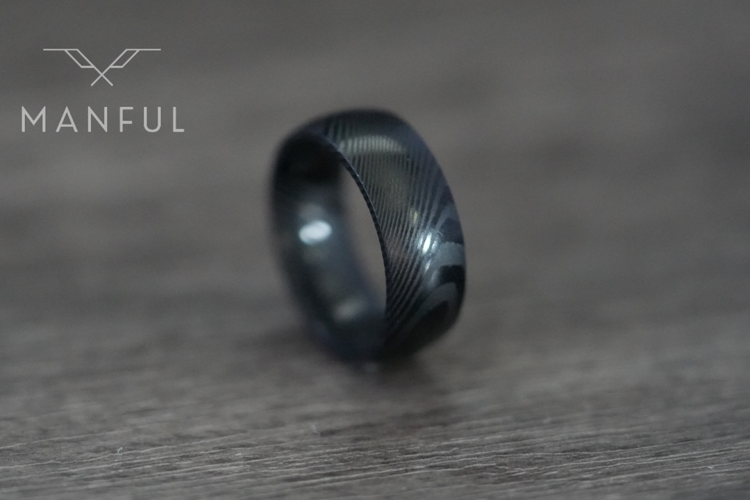 Damascus Steel Band Black, resting on its side on a wood surface with a blurred backdrop