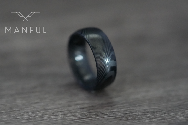 Damascus Steel Band Black, resting on its side on a wood surface with a blurred backdrop