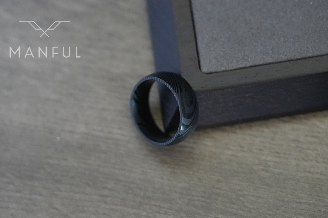 Damascus Steel Band Black, balanced on the corner of a dark box on a wood surface