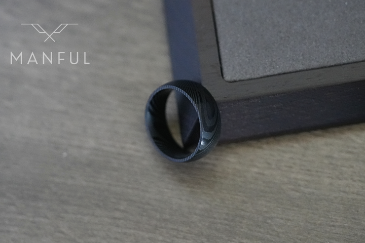 Damascus Steel Band Black, balanced on the corner of a dark box on a wood surface