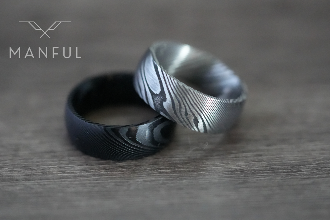 Damascus Steel Band Black, paired on wood beside a lighter silver damascus ring