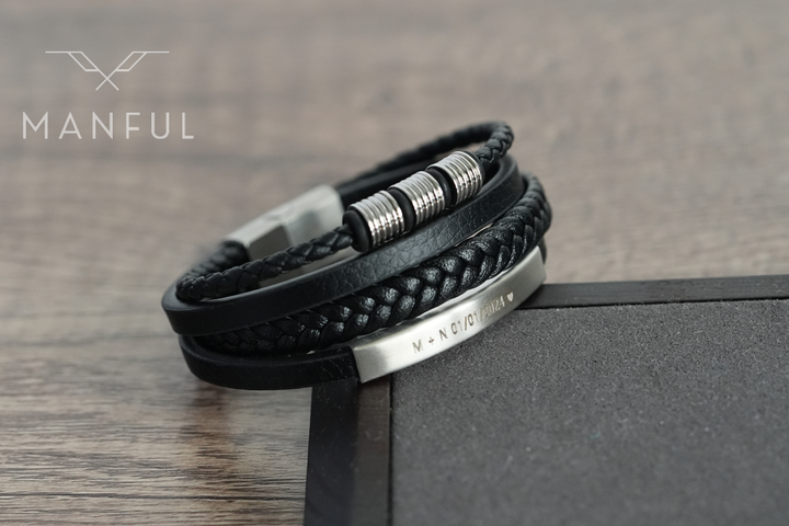 Black & Silver Leather Bracelet, engraved bar clasp draped over a dark box edge on wood