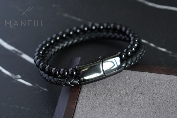 Onyx Bead And Leather Bracelet, coiled on dark marble beside a brown leather strip prop