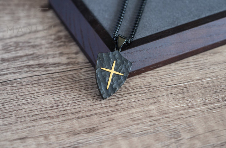 Shield Cross Pendant Black, resting on a striped wooden box with the chain draped across it