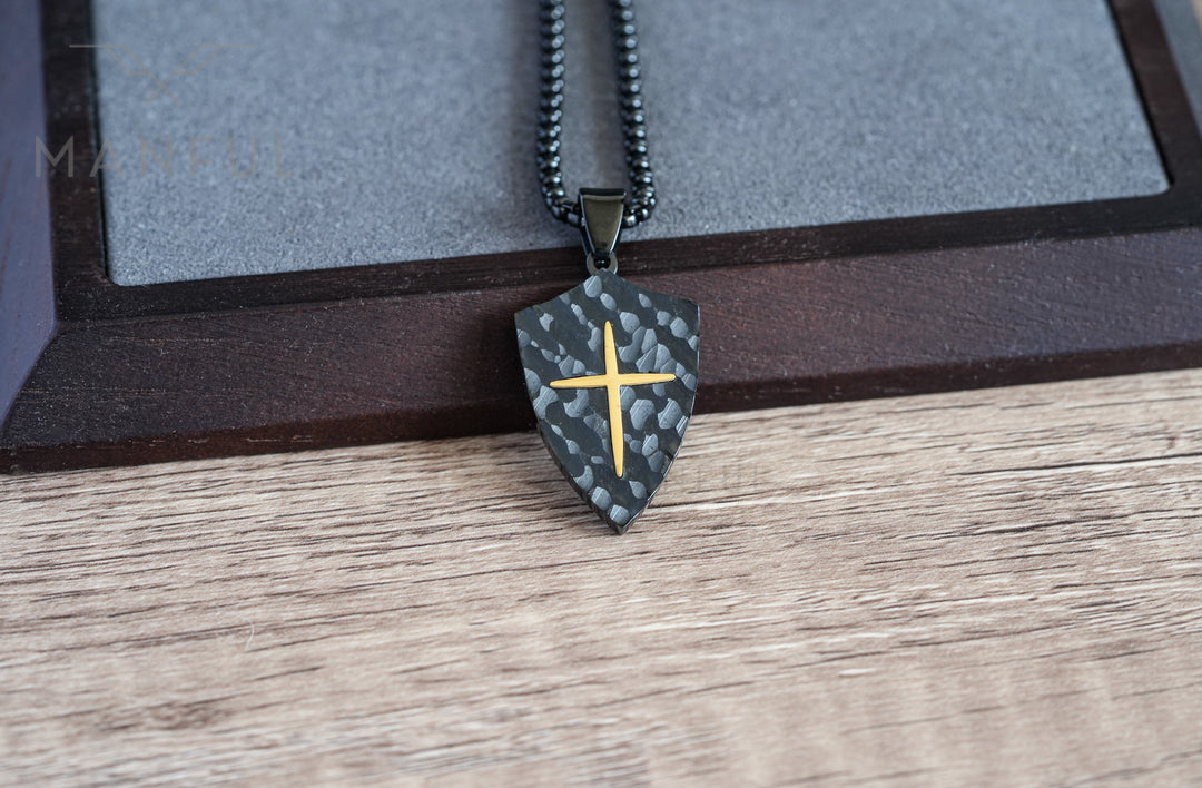 Shield Cross Pendant Black, hanging in front of a grey stone tile and dark wood block