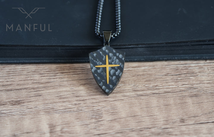 Shield Cross Pendant Black, resting against a black book on a wood table