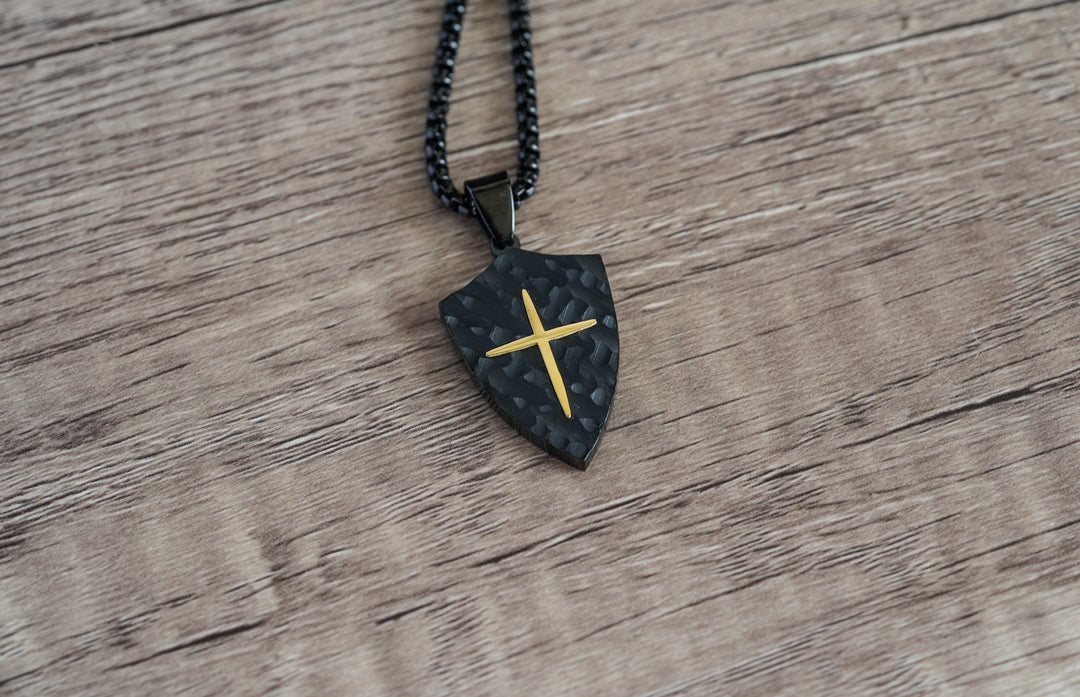 Shield Cross Pendant Black, a flat lay on a wood table with the chain coiled beside it