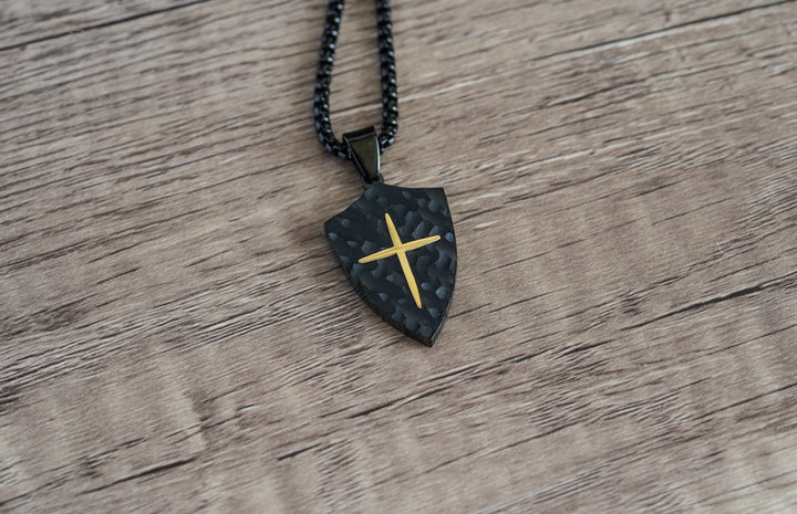 Shield Cross Pendant Black, a flat lay on a wood table with the chain coiled beside it