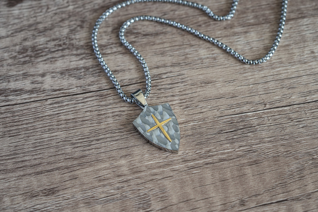 Shield Cross Pendant Silver, lying flat on a wood surface with the chain coiled beside it