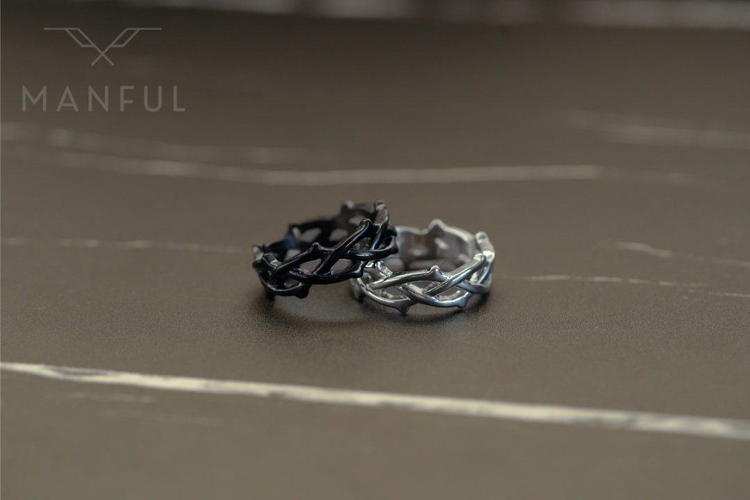 Thorn Ring Black, close-up beside the silver version resting together on a dark stone surface