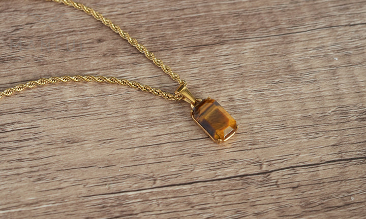 Tigers Eye Stone Pendant Gold, lying flat on a wood surface with the chain coiled beside it