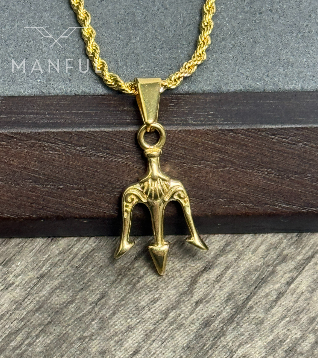 Trident Pendant Necklace Gold, resting against a wooden box on a stone surface